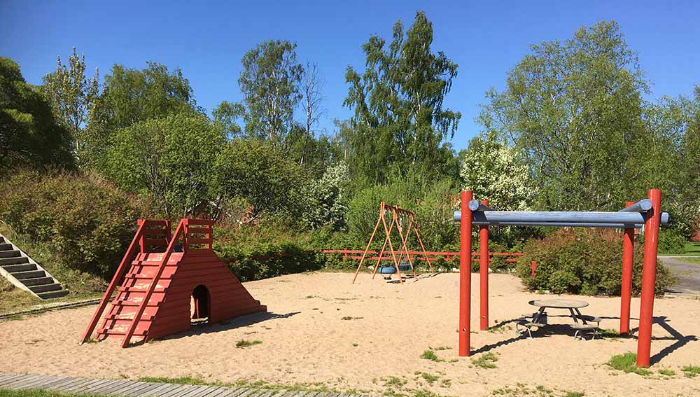 Klättertorn med rutschkana. En trapp upp på en gräskulle bredvid. Gungor i bakgrunden. Till höger en pergola eller klätterställning med bord och sittplatser under