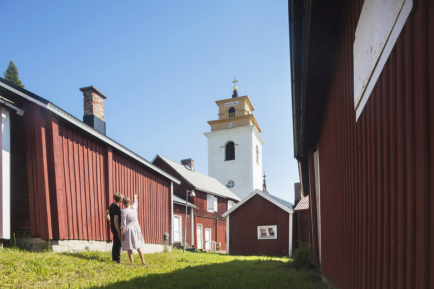 Två personer mellan röda kyrkstugor. Blå himmel och kyrktorn i bakgrunden.